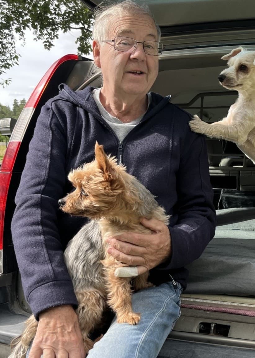 Äldre man sitter i bakluckan på sin bil med två små hundar.