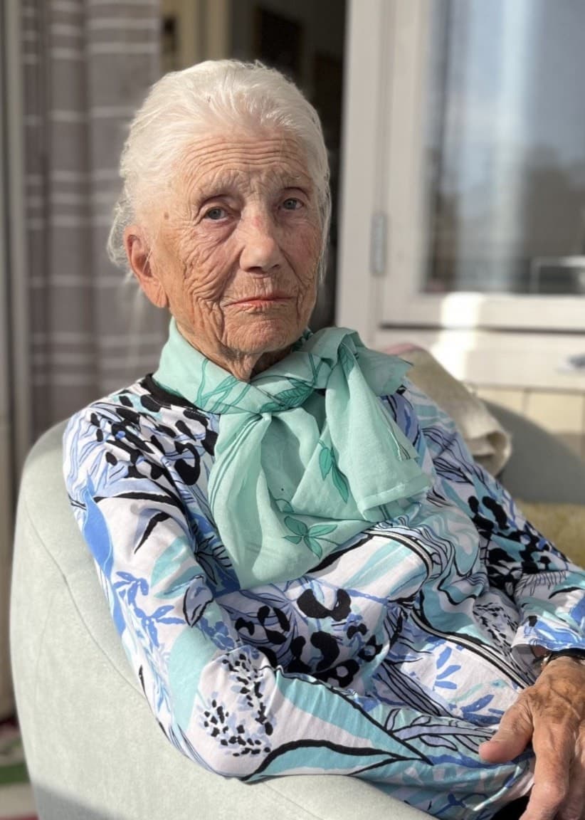 En äldre person sitter i en fåtölj på en solig balkong. Personen bär en mönstrad blus och en grön halsduk. Balkongen är inredd med en dörr, fönster och en färgglad matta.