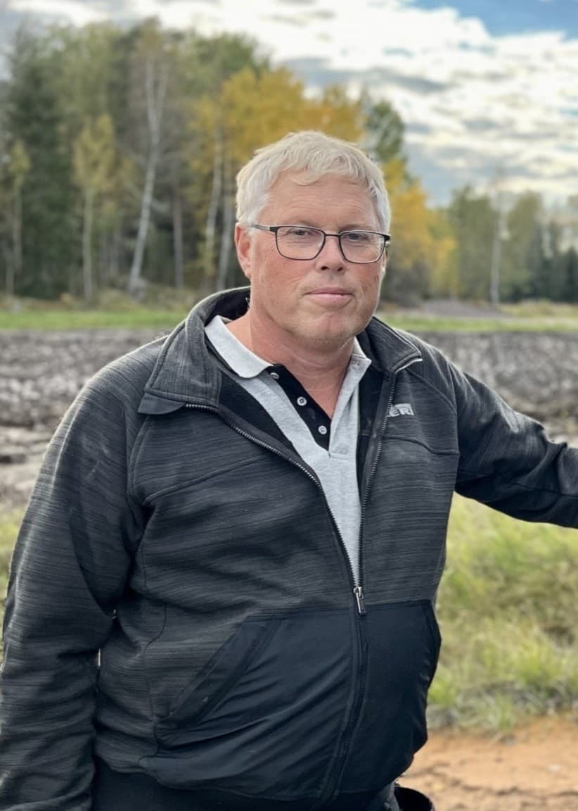 Morgan Jakobsson står vid två stora traktordäck på en landsbygd med ett plöjt fält i bakgrunden och ett hus omgiven av träd. Himlen är klar med några moln.