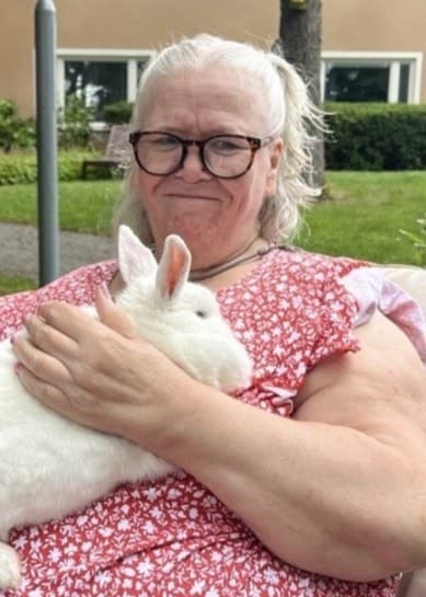 En äldre kvinna sitter i en rullstol iklädd en röd blommig klänning och håller en vit kanin i famnen. I bakgrunden syns en grönskande trädgård och ett bostadshus.