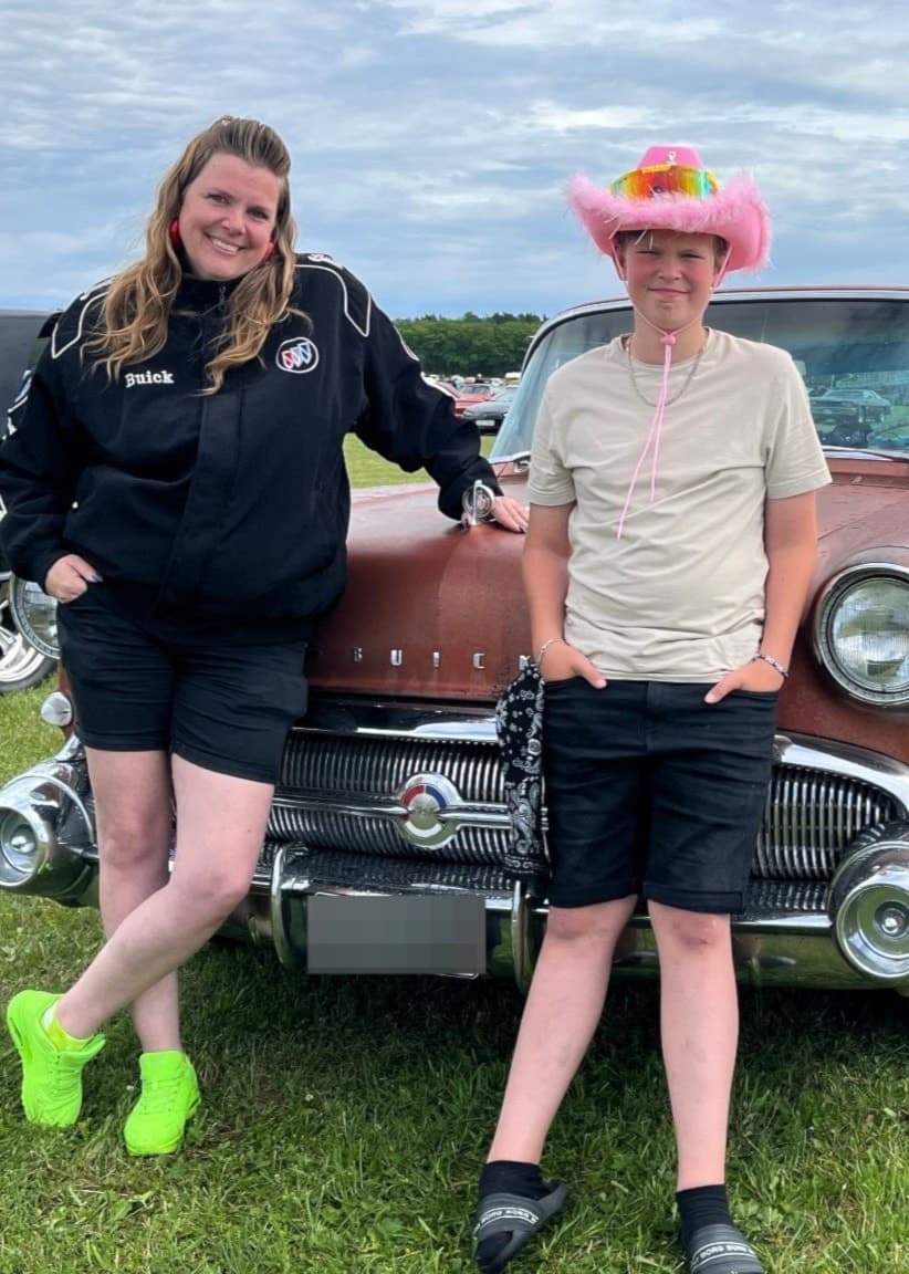 Anna Karlberg står bredvid 11-åriga Loke framför en klassisk bil på en bilträff. Loke bär en rosa cowboyhatt och ler. Gräsmatta och fler bilar syns i bakgrunden under en molnig himmel.