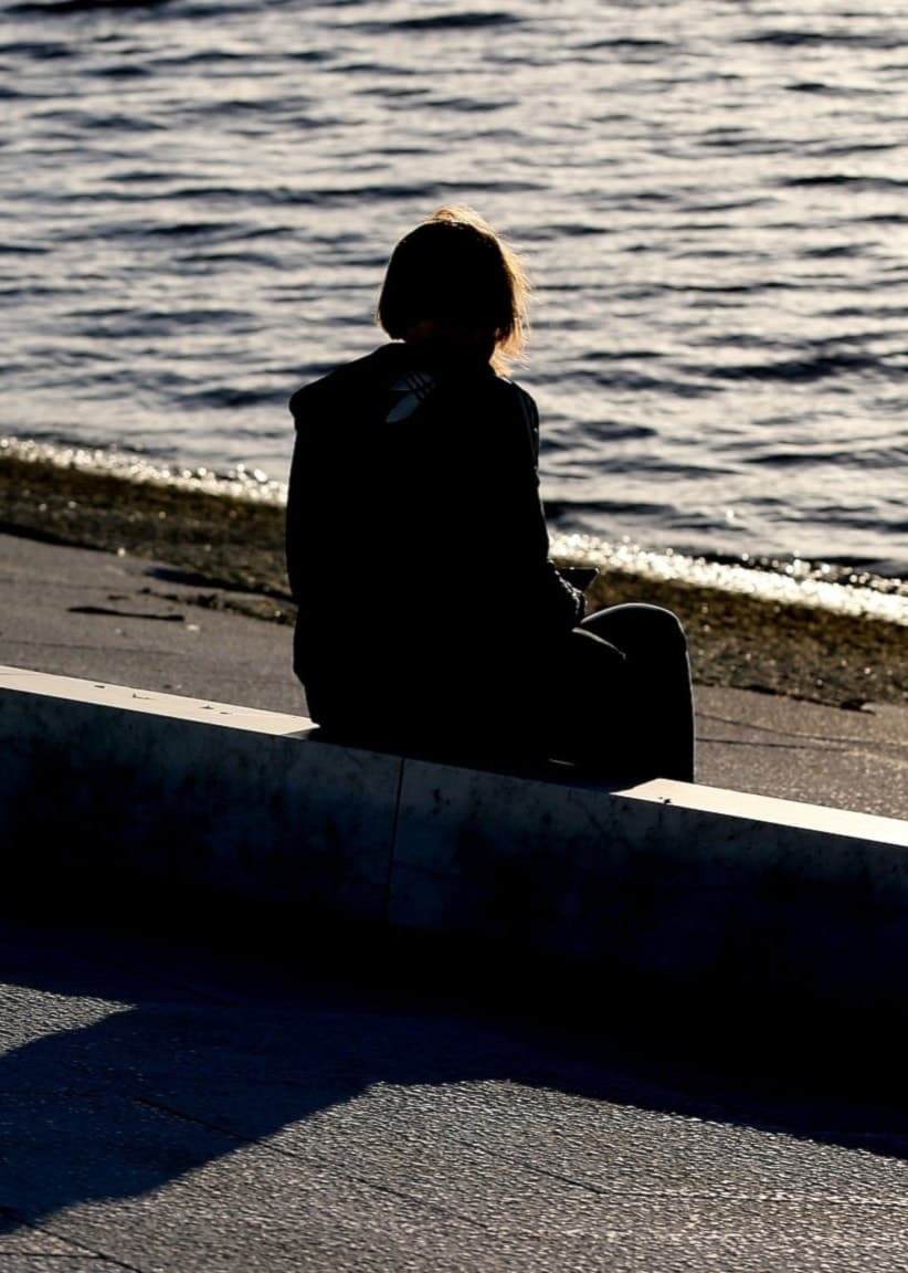 En ensam person sitter vid vattnet på en stenmur i motljus. Skuggor och vågor skapar en melankolisk stämning. Personen är vänd bort från kameran och omgiven av ett stilla landskap.