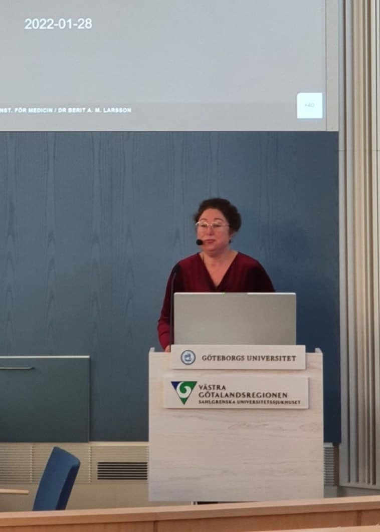 Berit Larsson står vid ett podie, med mick och en presentation uppe på en digital skärm i bakgrunden,  inuti en byggnad vid Göteborgs Universitet. 