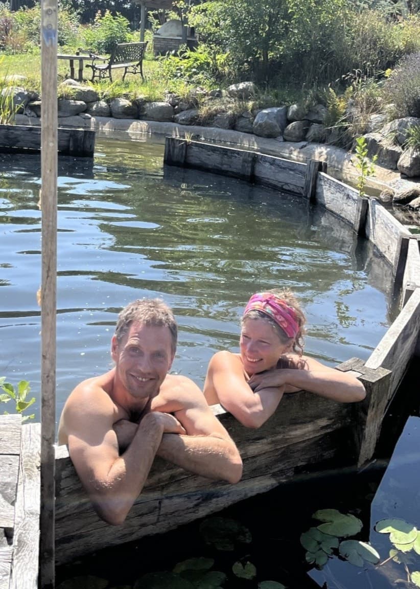 Etta och Jona kopplar av i en naturlig ekopool omgiven av grönska, träbrygga och näckrosor. Miljön skapar en lugn och harmonisk atmosfär med fokus på hållbarhet och natur.
