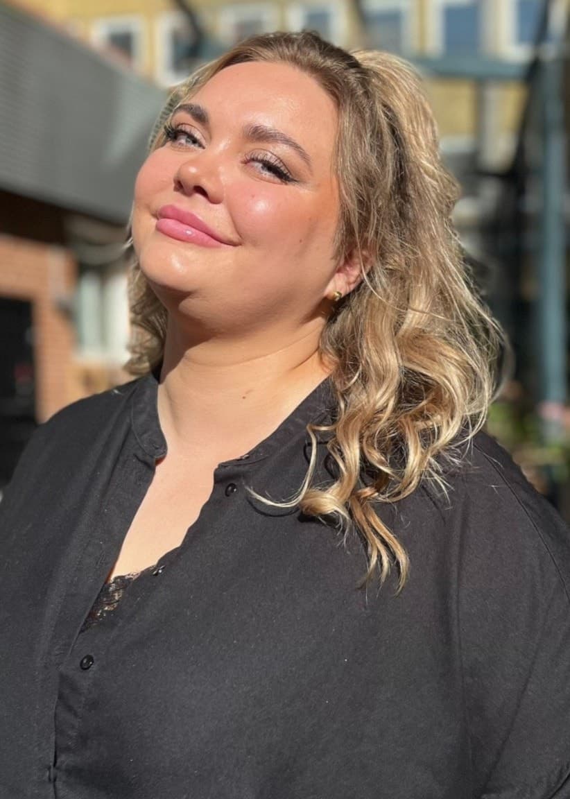 Newgirl, finalist i P4 Nästa Stjärna, står utomhus på en innergård med solbelysta byggnader och grönska i bakgrunden.