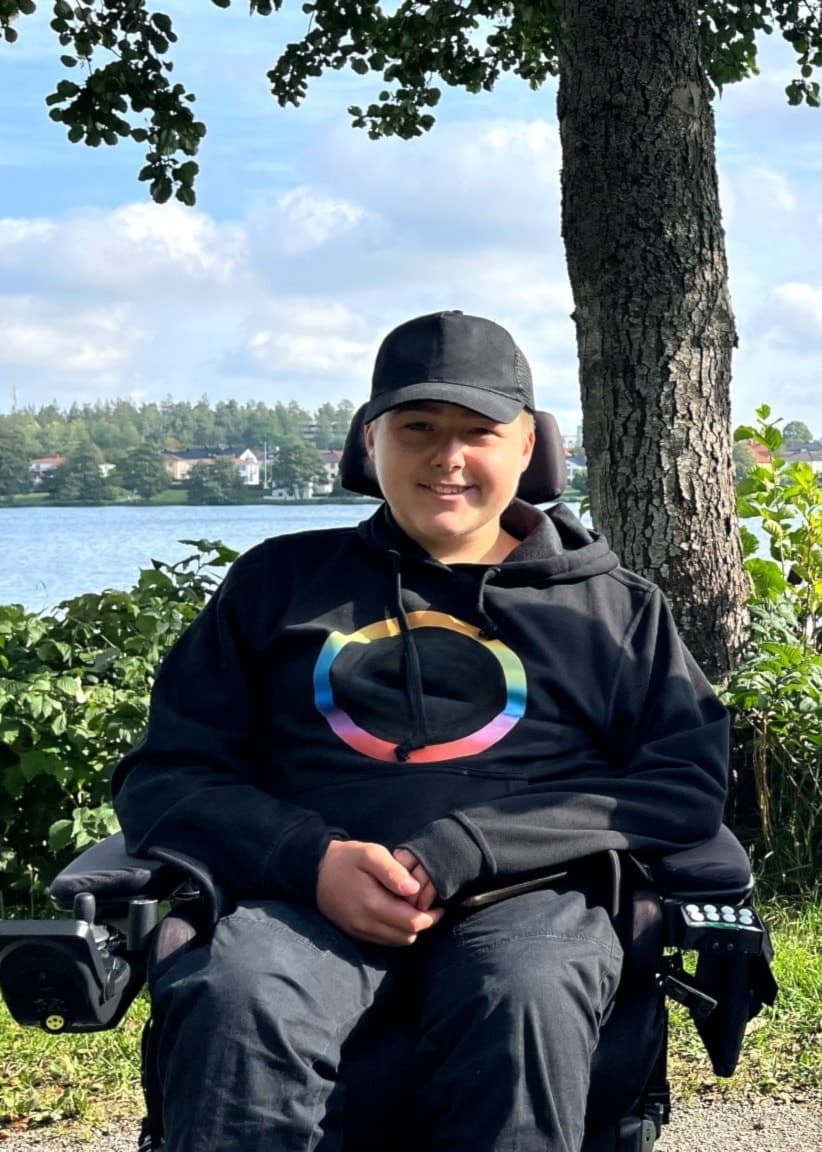 Melwin Stålhammar och Agnetha Hugo sitter utomhus i sina rullstolar vid en sjö med grönska och blå himmel i bakgrunden, en scen som förmedlar lugn och naturskönhet.
