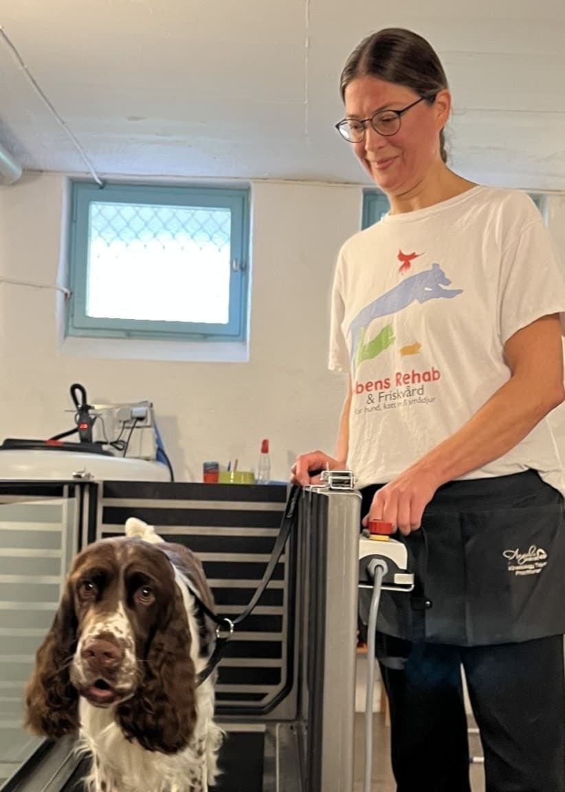 Jenny Bertilsson står i en lokal med ljusa väggar och håller i hunden Foppa, som befinner sig på ett löpband anpassat för hundar. Miljön innehåller träningsutrustning och en grön stol.
