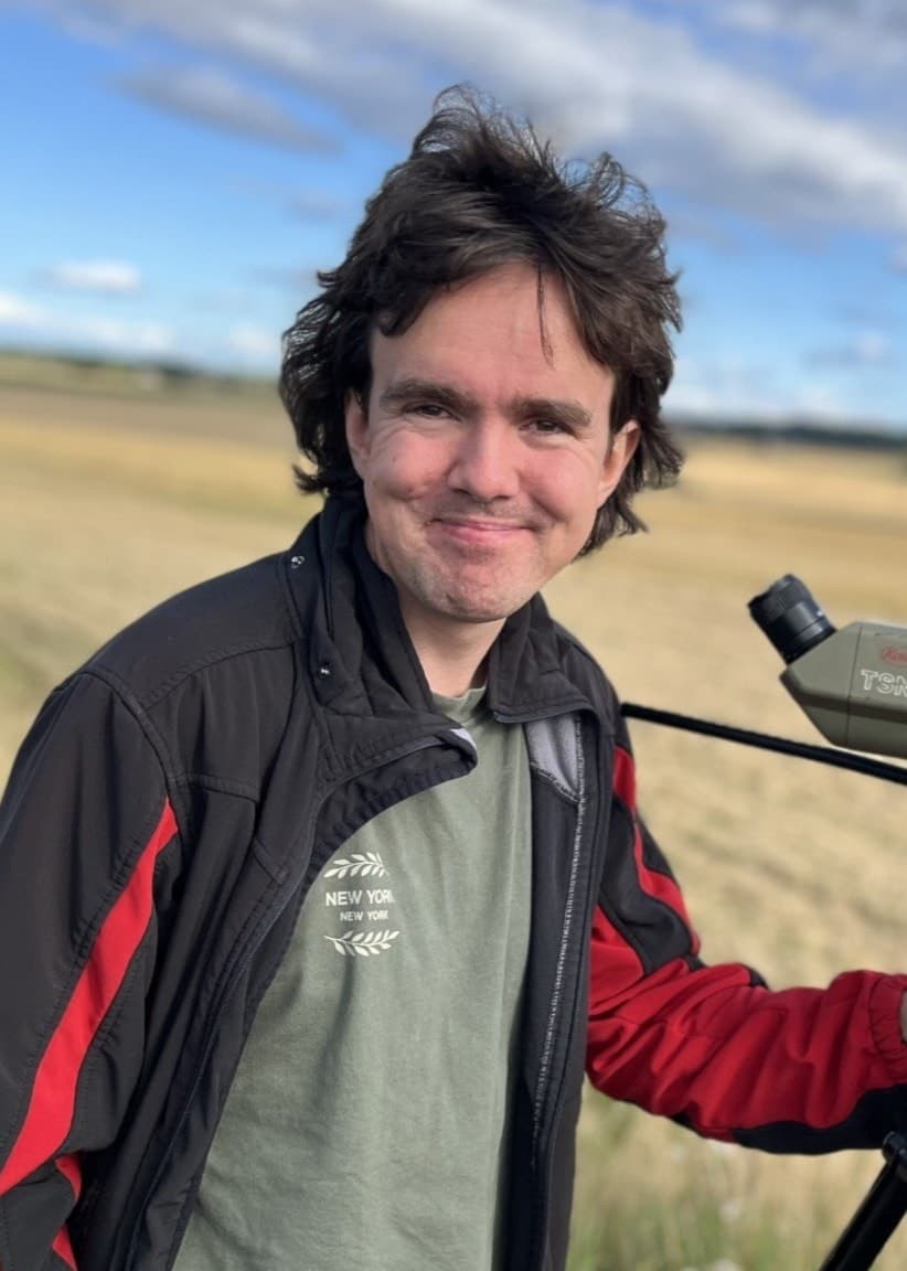 Leo Rudberg, fågelskådare, står på en åker med en kikare monterad på ett stativ. Landskapet är öppet med blå himmel och några vita moln i bakgrunden.