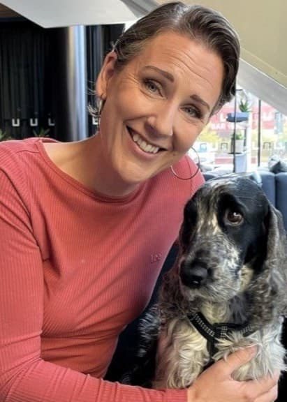 Katarina Häll sitter på en soffa i ett modernt vardagsrum med en hund i knät. Bakgrunden visar stora fönster, grå gardiner och en grön växt till höger.