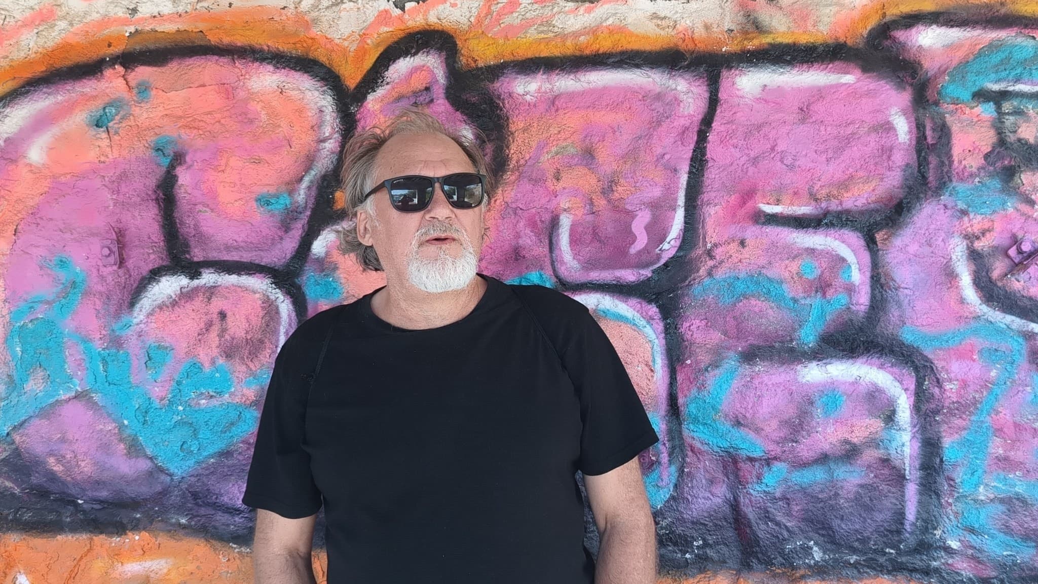man med grått hår, svart t-shirt och solglasögon står framför färgglad graffitivägg