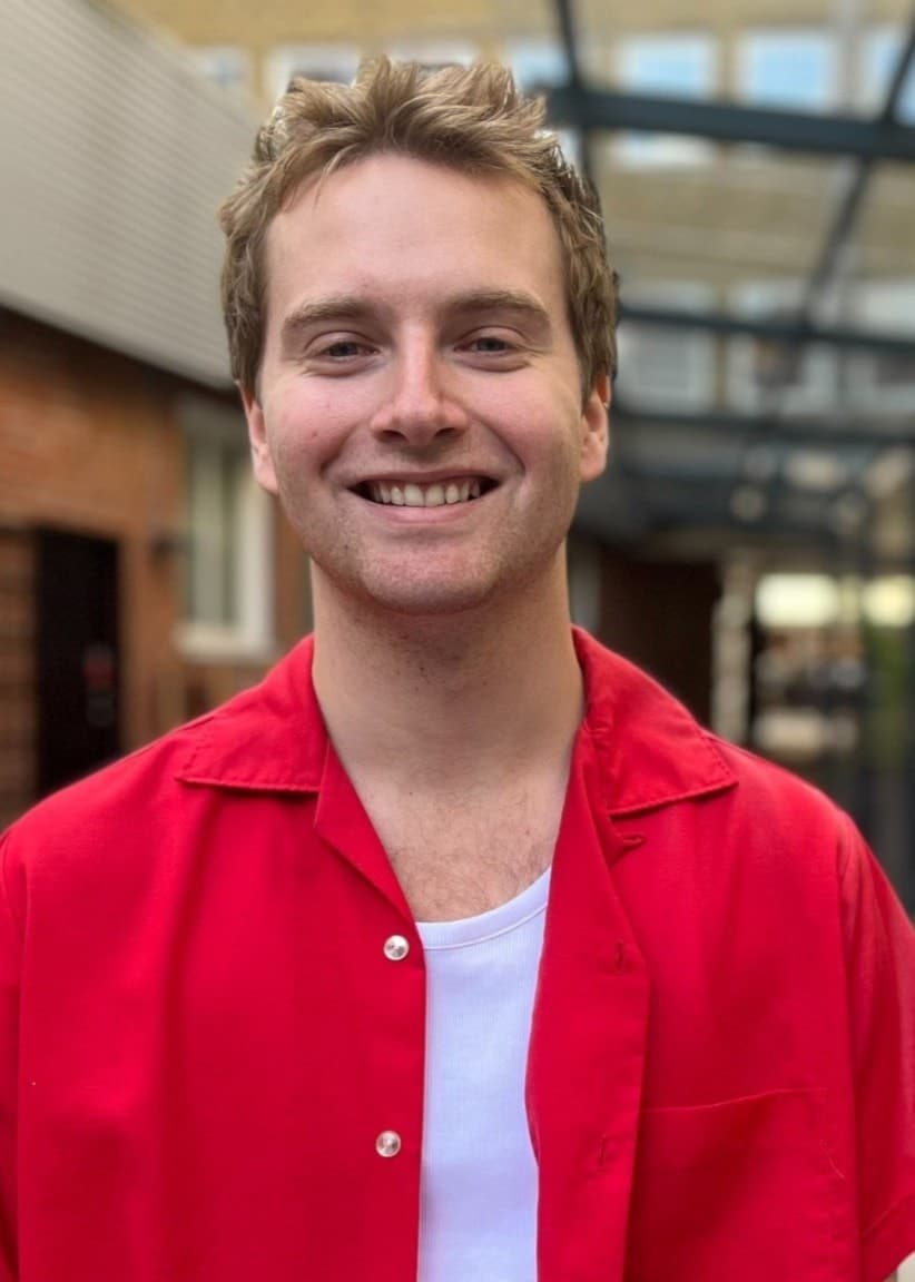Jakob Willfors, finalist i P4 Nästa Stjärna Malmöhus, står utomhus i en röd skjorta framför en modern byggnad med glasdetaljer och växtprydd gång.
