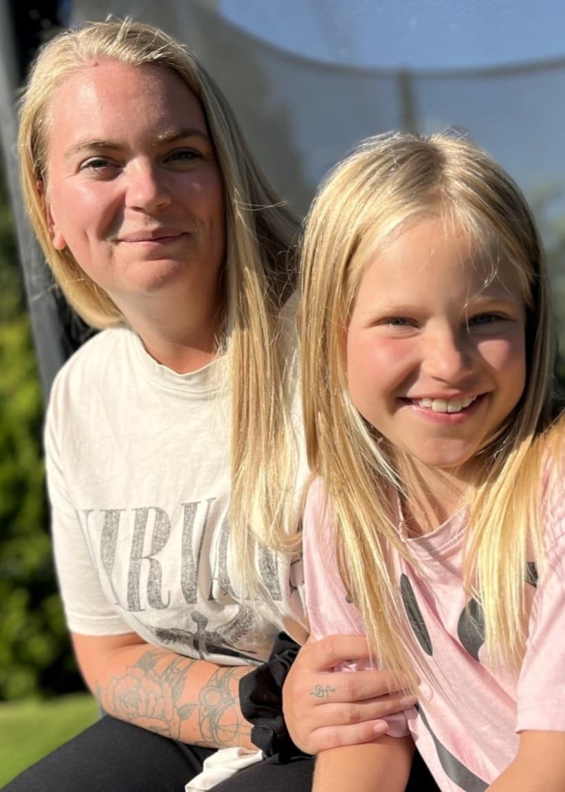 Ronja Luff och hennes dotter Juni sitter tillsammans utomhus på en studsmatta i solskenet. Grönskande buskar och blå himmel syns i bakgrunden. Juni har återhämtat sig efter en sjukdomsperiod.