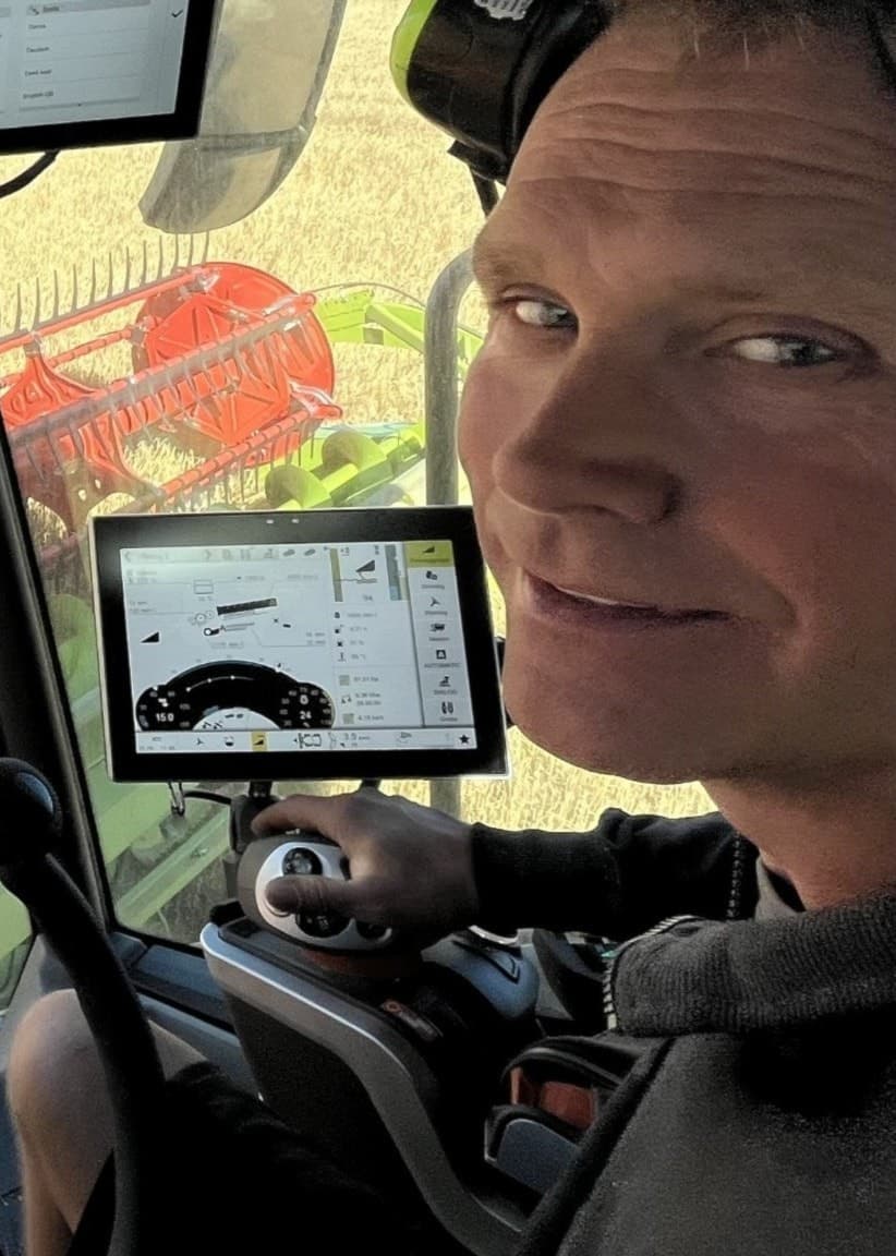En skördetröska sedd inifrån förarhytten, med digitala skärmar som visar data. Utanför syns ett stort fält med spannmål som skördas.