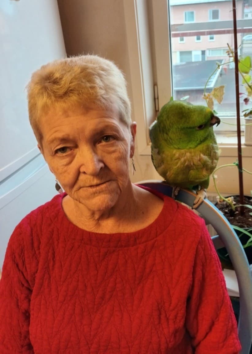 En person med röd tröja sitter inomhus med en grön papegoja på axeln, omgiven av krukväxter. Bredvid syns en solig havsutsikt med solstolar och parasoller vid en terrass.
