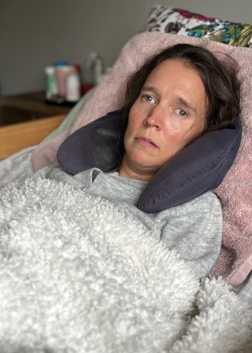 En person ligger i en säng med en ljusgrå filt över sig och huvudet vilande på en kudde. I bakgrunden syns en del av ett kök och en stol.