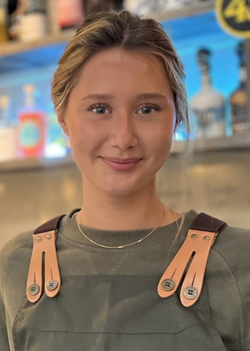 Alice Hamlund, klädd i arbetsförkläde, står vid en bardisk med flaskor och tappkranar i bakgrunden. Hon delar sin upplevelse som sommarjobbare inom serviceyrket.