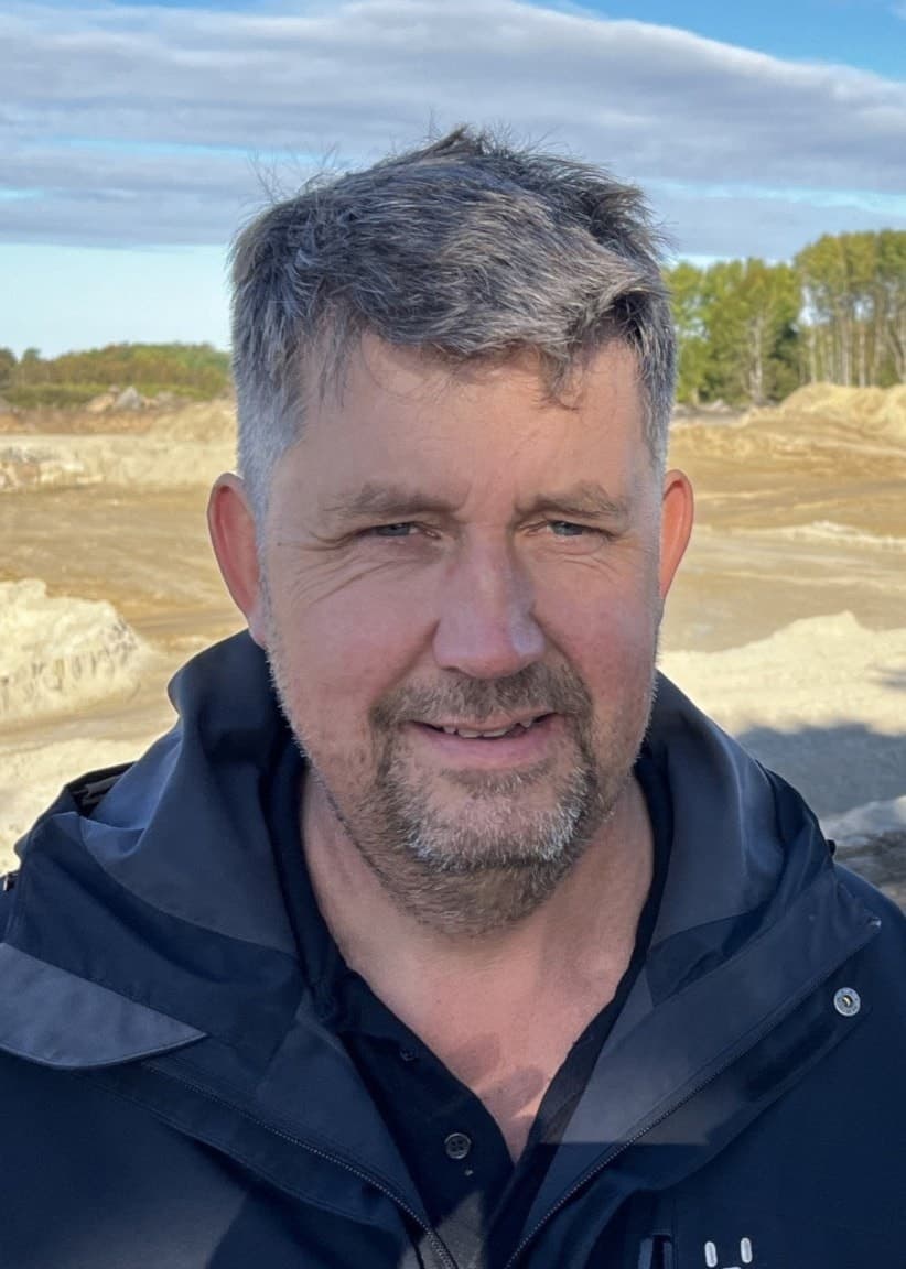 En man i blå jacka står framför en byggarbetsplats där fler arbetsfordon syns i bakgrunden.