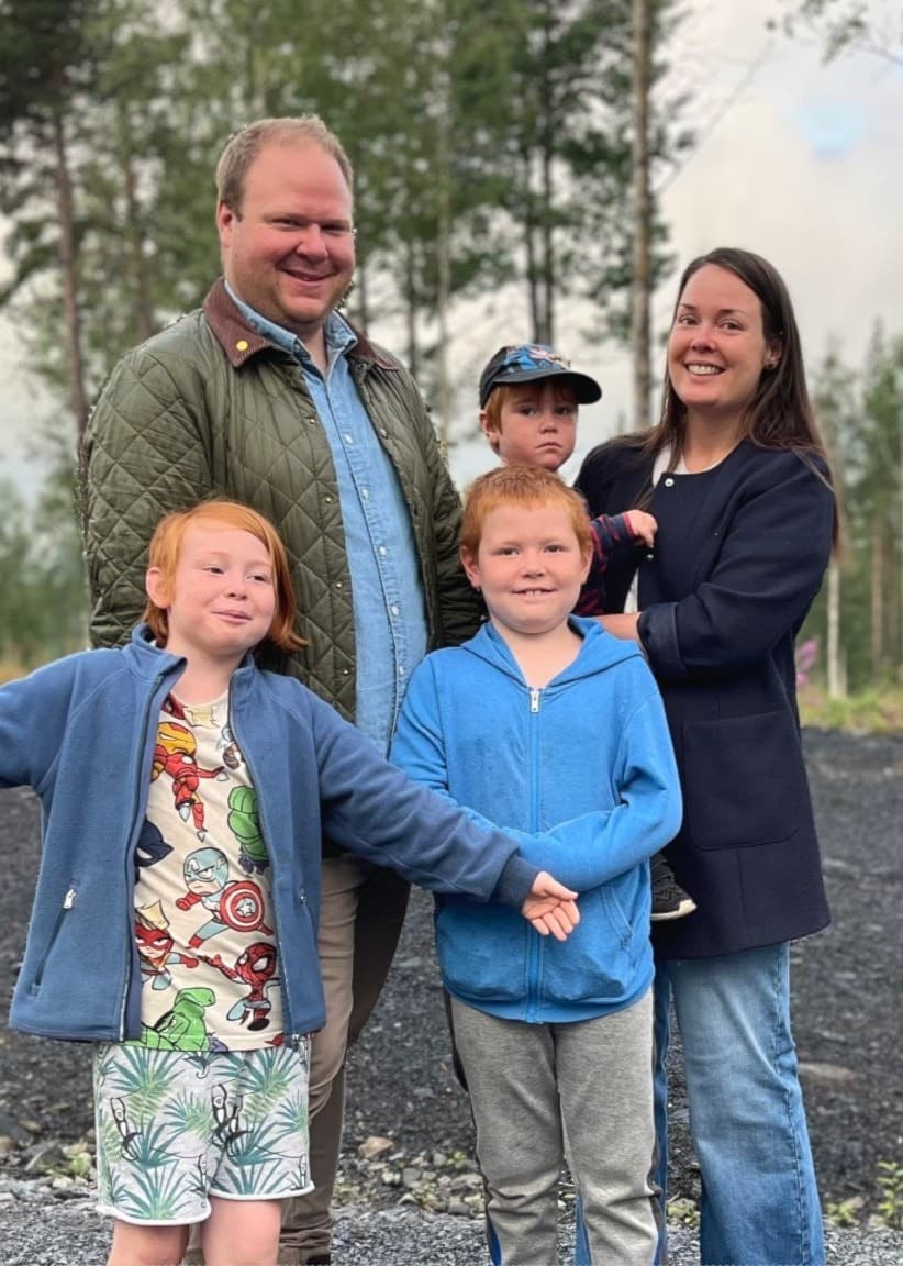 Mattias och Linnea Barkenfelt står med sina barn på en grusväg omgiven av skog. Familjen ler och barnen ser glada ut, med höga träd och en molnig himmel i bakgrunden.