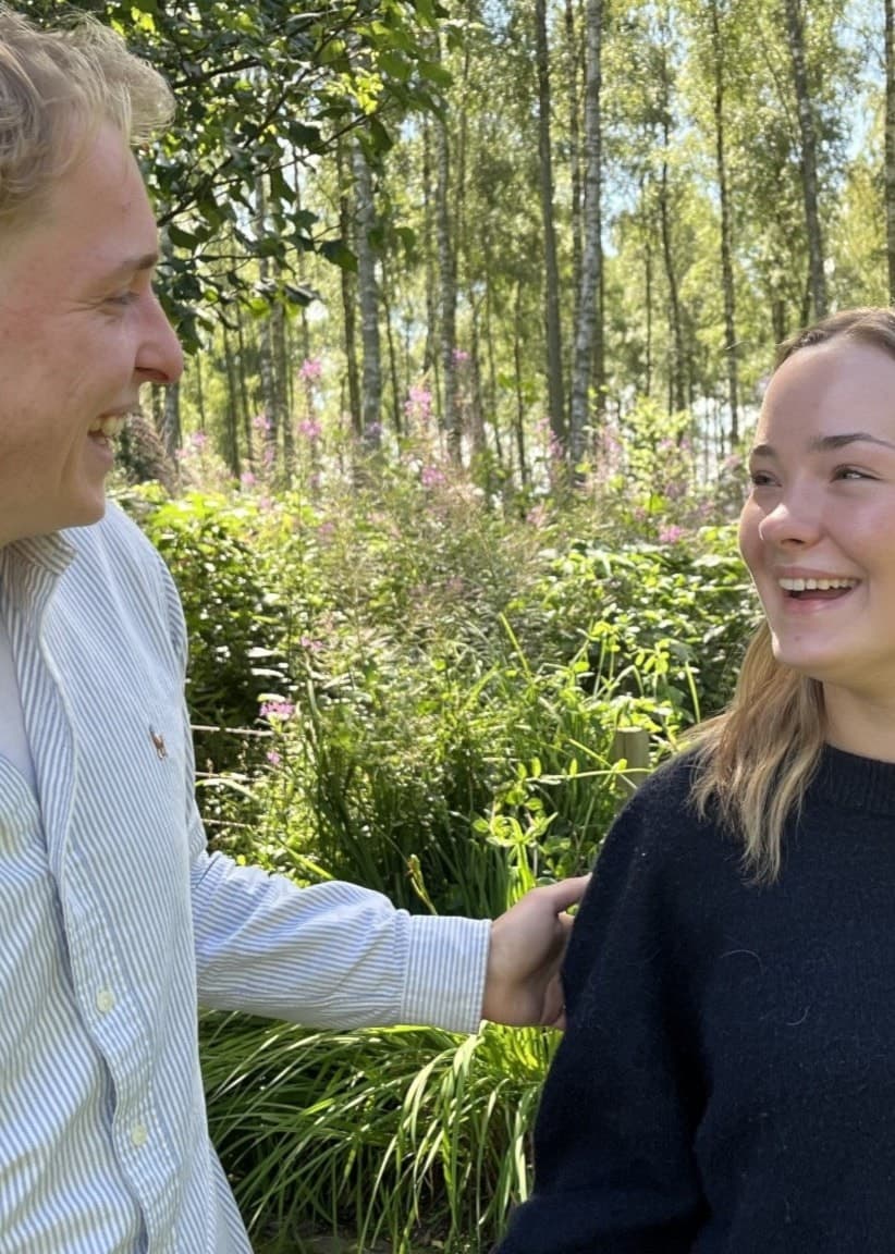 Liam Karlsson och Linnéa Ljung står utomhus i en grönskande miljö med träd och buskar i bakgrunden. Liam bär en ljus skjorta och Linnéa en mörk tröja, och de tittar på varandra och ler.