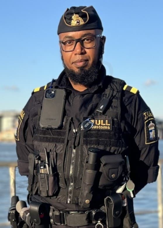 En polis står vid kajen i Göteborg med utsikt över vattnet och den ikoniska skyskrapan Karlatornet i bakgrunden. Solen skiner och byggnader samt kranar syns på andra sidan vattnet.