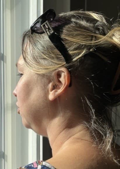 Kvinna med blont hår uppsatt i en knut står vid ett fönster och tittar ut. Solljus lyser in genom fönstret och träffar hennes axel. Solglasögon vilar på hennes huvud.