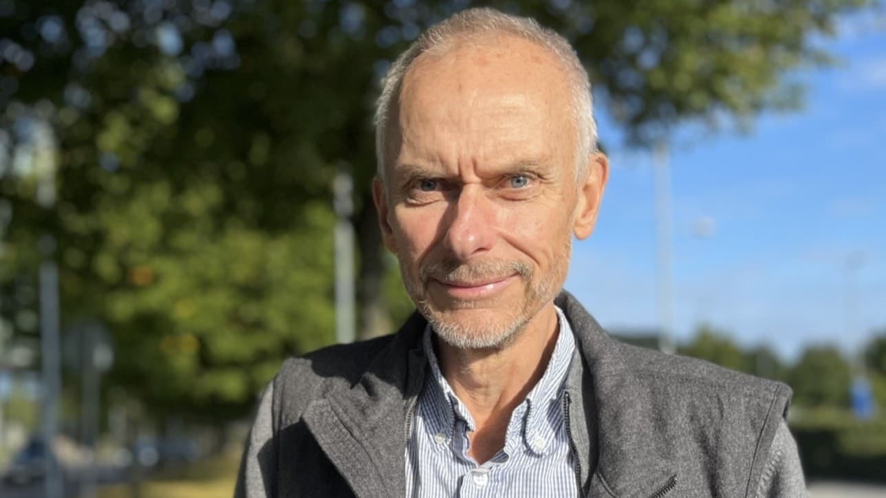 Halvbild på en man i skjorta och grå väst som kisar mot solen. I bakgrunden syns träden mörkgrå vägg.