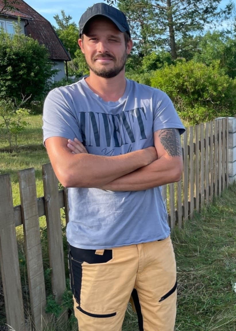 En man står med armarna i kors vid en landsväg nära en busshållplats. Han bär en grå t-shirt och beige shorts. Omgivningen består av grönska, ett trästaket och ett bostadshus i bakgrunden.
