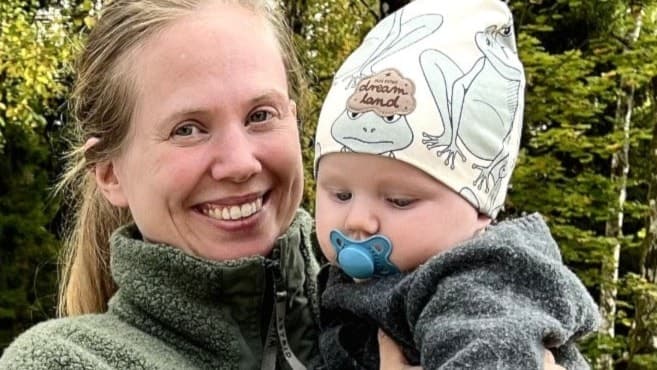 Tvåbarnsmamman Ida Hermansson står utomhus med sitt barn, Folke 5 månader, på armen, omgiven av grönska. Framför henne syns en barnvagn på en grusväg i en naturskön miljö med träd i bakgrunden.