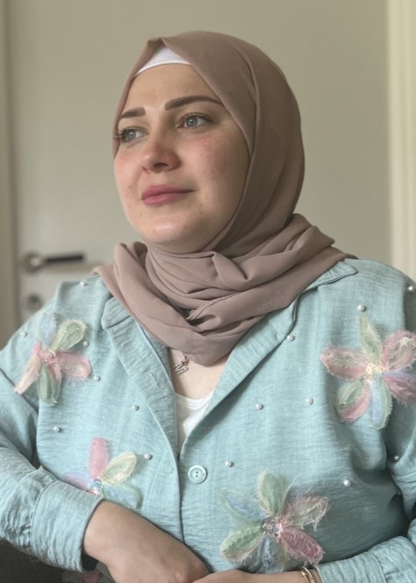 Marwa Karkanawi sitter vid ett bord med en flätad bordstablett framför sig. Hon bär en turkos hijab och en ljusblå blus dekorerad med blommotiv. Miljön är ljus och minimalistisk. Bild på entén till Risbergska.