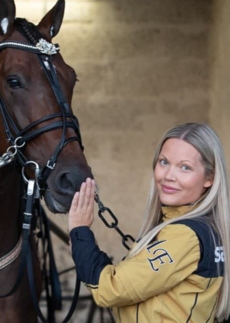 Michelle Eriksson klädd i gul jacka står i ett stall på Kalmartravet och interagerar med en brun travhäst utrustad med sele, omgiven av hästutrustning.