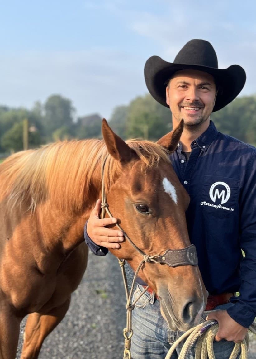 Man i cowboyhatt, står med en häst framför en hage