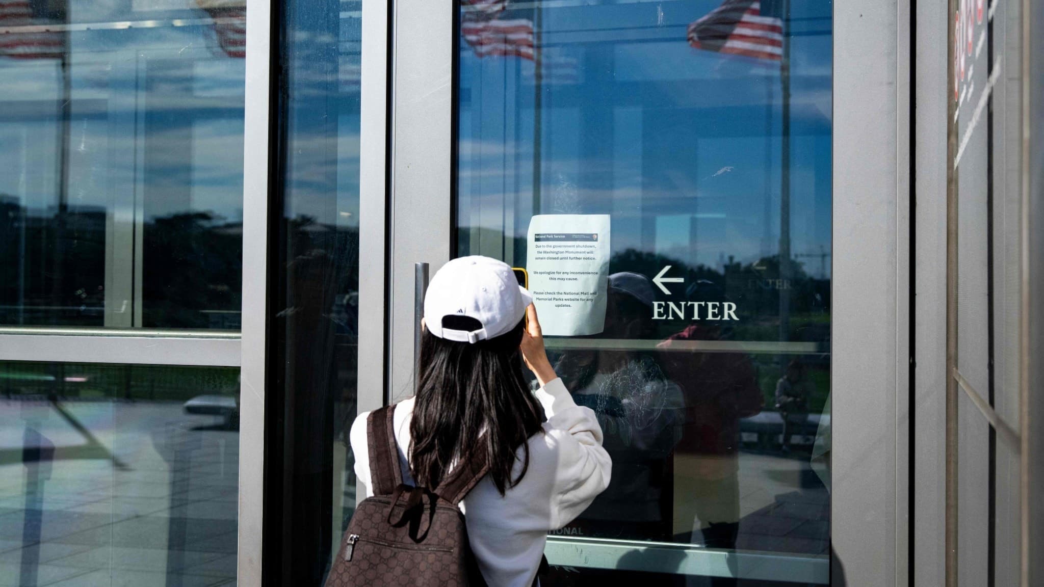 En kvinna fotograferar en skylt på Washington Monument som visar att det är stängt på grund av en regeringsnedstängning. Amerikanska flaggor reflekteras i byggnadens glasfasad.