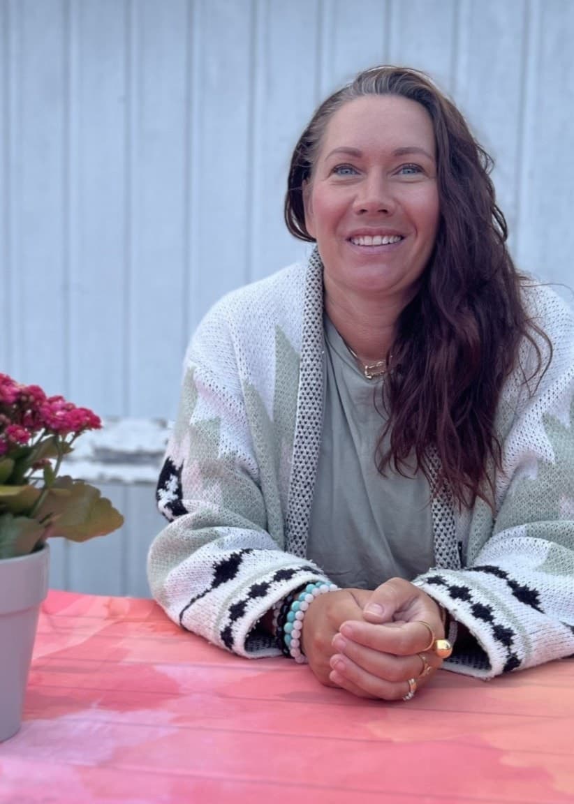 En person sitter vid ett bord täckt av en färgstark duk, med en blomkruka med röda blommor framför sig. Bakgrunden visar en vit trävägg och en trädgård med staket.