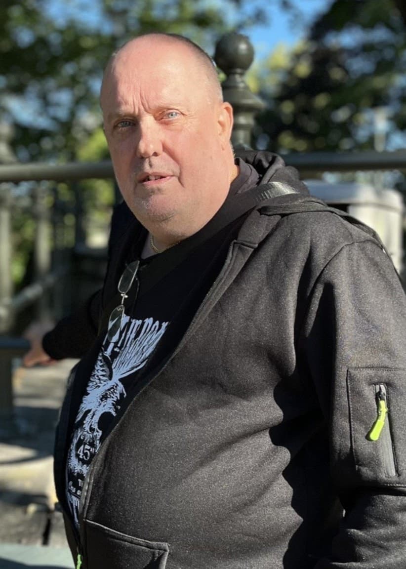 Peter Glans från Borås i svart tröja står utomhus. Bredvid syns en hand som håller en öppnad cachegömma, använd vid geocaching, med ett lila föremål inuti.