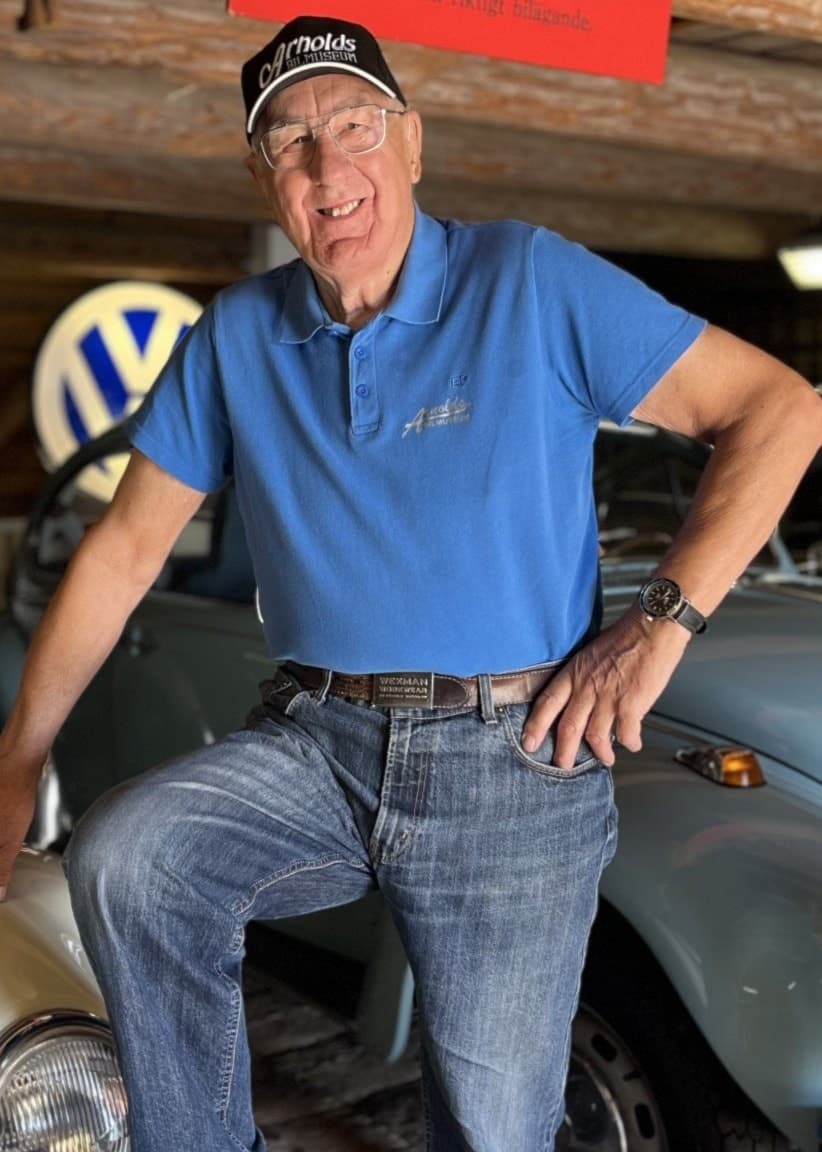 Arnold Sundström poserar bredvid en vit veteranbil i ett museum med träinteriör, omgiven av ytterligare klassiska bilar och nostalgiska skyltar.