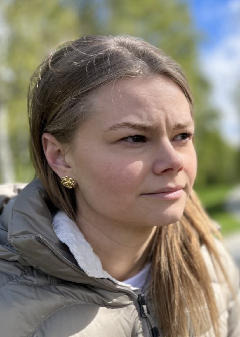 Linn Gustafsson ses utomhus i en park med grönskande träd och gräs i bakgrunden, iklädd en beige jacka. Solen skiner och himlen är delvis molnig.