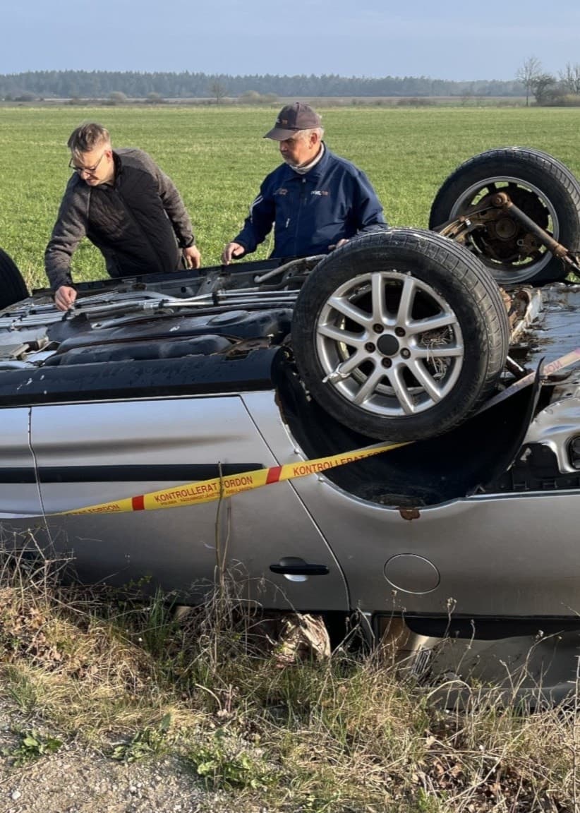 En silverfärgad bil ligger upp och ned i ett dike med varseltejp runtom, två män står och inspekterar bilen. 