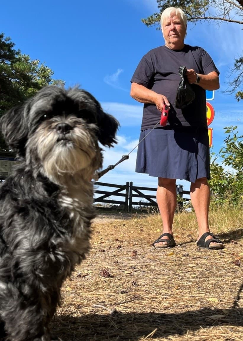 En äldre man står utomhus i en trädgård med två soptunnor i bakgrunden. En liten svartvit hund sitter i förgrunden på en torr gräsmatta under klarblå himmel.