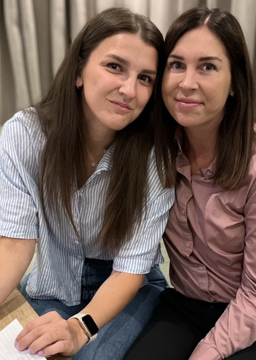 Johanna Larsson och Emelie Hagström sitter bredvid varandra i en vardagsrumsmiljö. På bordet framför dem ligger ett anteckningsblock och en penna.