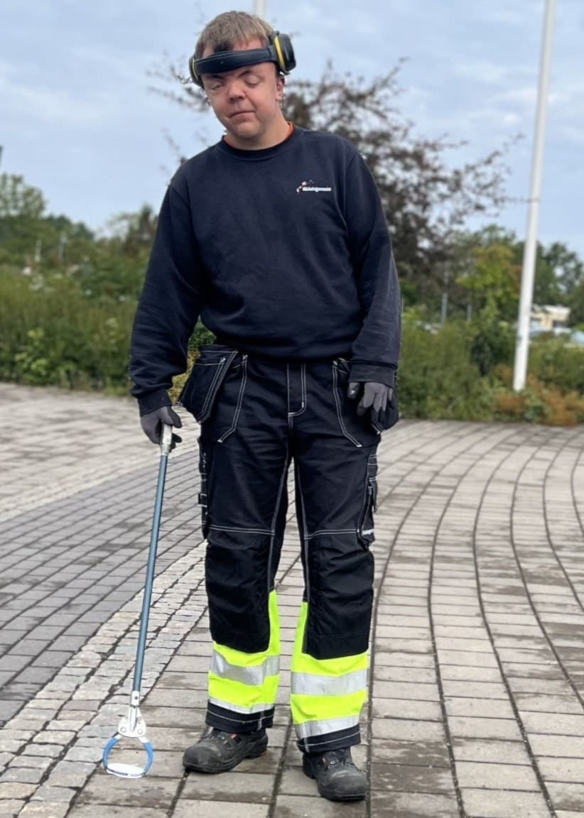 Måns Pettersson står utomhus på en stenlagd yta, klädd i arbetskläder med reflexdetaljer. Han bär skyddsglasögon och håller i verktyg, omgiven av träd och cykelställ i bakgrunden.