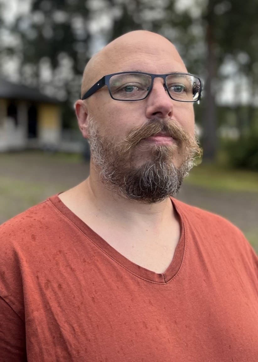 Per Björnholm står utomhus framför en gul byggnad med skog i bakgrunden. Han bär en röd tröja och omgivningen är molnig och grå.