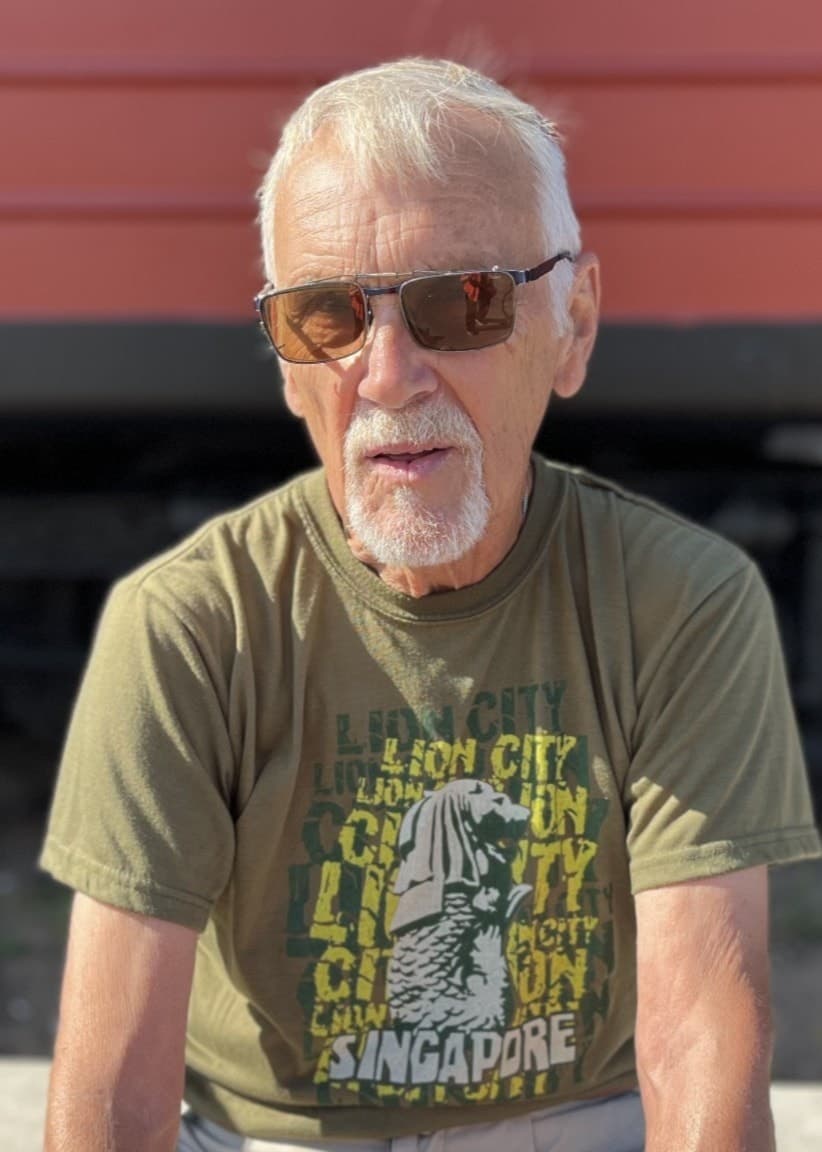 83-årige Egon Kristersson sitter vid ett bord utomhus, klädd i en grön t-shirt, med en tågvagn i bakgrunden. Han tränar regelbundet för att hålla kroppen aktiv och nå sitt mål.