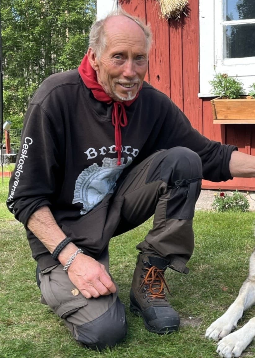 En man i svart tröja sitter på huk bredvid en hund, Bronco, framför ett rött trähus med vita fönster i en lantlig miljö.