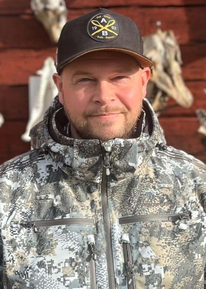 Björn Taavo står framför en trävägg dekorerad med älghorn och djurhudar, klädd i kamouflagemönstrad jacka och keps, i samband med att han använder lockrop för att locka älgar.