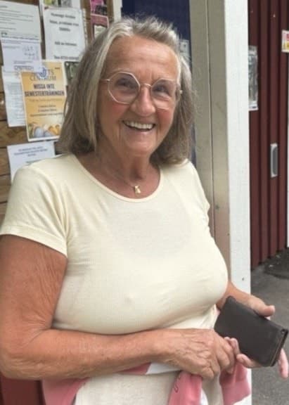Eva Syrén står utanför en röd byggnad vid Östra stranden, nära en toalett, och håller i en mobiltelefon. Bakom henne syns en anslagstavla med olika meddelanden.