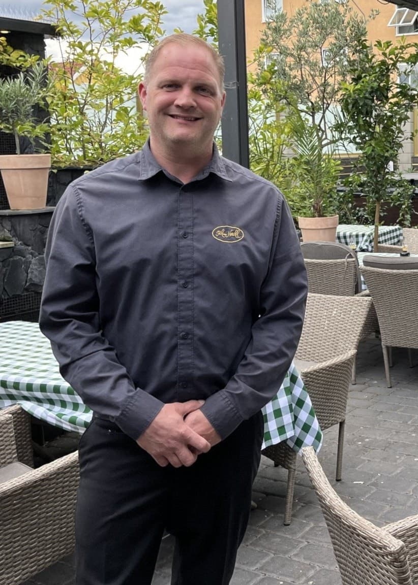 Christian Ryning, ägare av Hotell Palace, står på en grönskande uteservering med rutiga dukar och rottingmöbler, omgiven av växter och bostadshus i bakgrunden. 