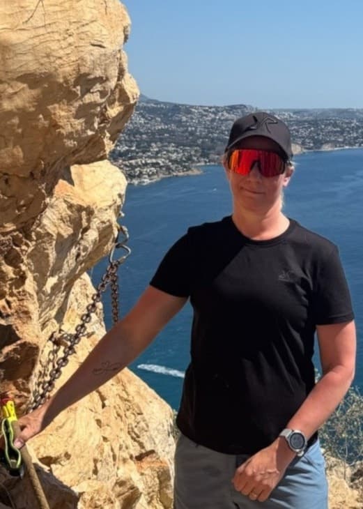 En person i svarta kläder och keps står vid en klippvägg med utsikt över en blå havsvik och en kuststad i bakgrunden. Soligt väder och klarblå himmel skapar en naturskön vy.