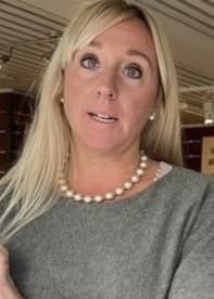 En kvinna med långt blont hår, klädd i en grå tröja och pärlhalsband, står i en butik fylld med inredningsdetaljer och dekorativa föremål.