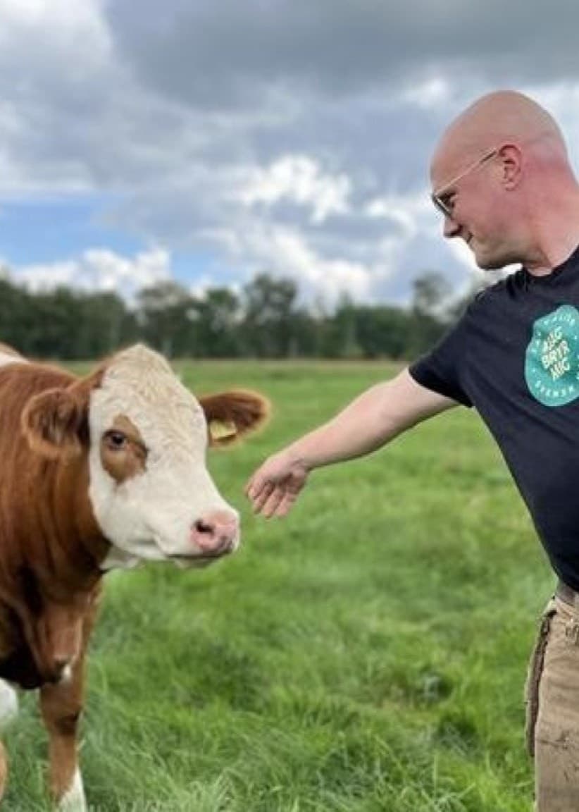 Henrik Gudmundsson, lantbrukare och vice ordförande i Lantbrukarnas Riksförbund i Skåne, står på en grön äng och interagerar med en ko. Solen lyser och moln syns på himlen.