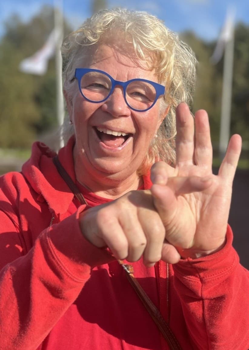 En kvinna i röd jacka står utomhus och gör en gest med händerna som symboliserar penetrationssex framför en byggnad och flaggstänger, omgiven av grönska och en klar himmel. Bygdens trick: Lämna barn på dagis – så ni kan göra syskon